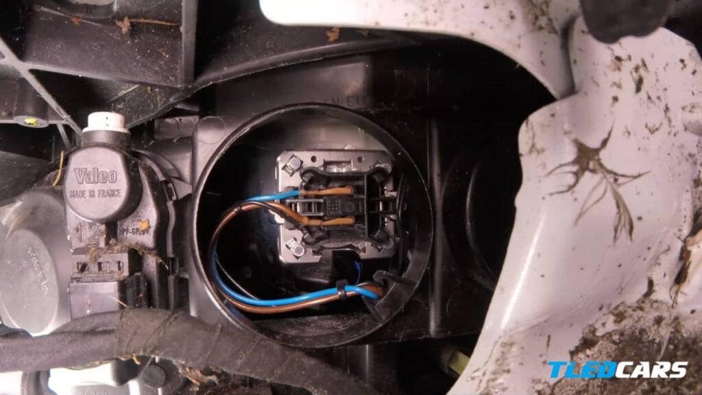 Gros plan détaillé du ressort métallique de fixation d'une ampoule H7 dans l'optique d'une Clio 3, montrant le point de pression pour débloquer l'ampoule sans casser le support.